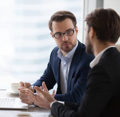 Two serious caucasian men in suits discussing or planning business issues in the office. Colleagues or client and consultant are sitting at the table next to each other and talking.